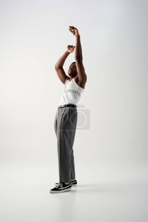 Full length of trendy afroamerican man in sleeveless t-shirt and pants raising hands and standing on grey background, contemporary shoot featuring stylish attire