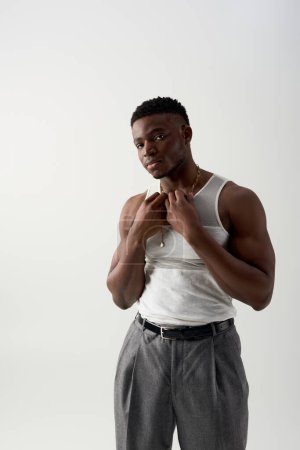 Muscular and young african american man in sleeveless t-shirt and pants touching necklace and looking at camera isolated on grey, contemporary shoot featuring casual attire