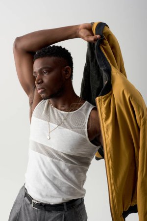 Muskulöses afrikanisch-amerikanisches männliches Model in Tank-Top mit Bomberjacke, das isoliert auf grauem, zeitgenössischem Shooting mit stylischer, lässiger Kleidung wegschaut, modisches Statement 