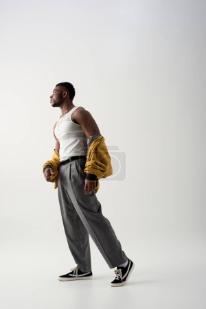 Téléchargez les photos : Vue latérale de l'homme afro-américain musclé en veste de bombardier et t-shirt sans manches debout sur fond gris, prise de vue contemporaine avec tenue élégante, déclaration de mode - en image libre de droit