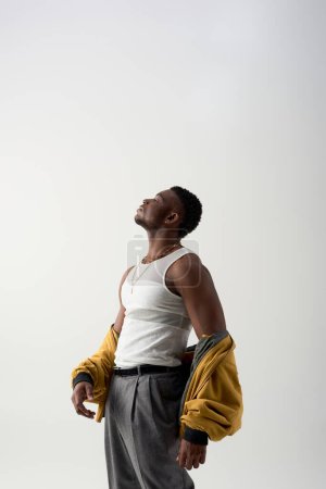 Photo for Side view of strong and trendy young african american man in tank top and bomber jacket standing isolated on grey, contemporary shoot featuring stylish attire, muscular - Royalty Free Image