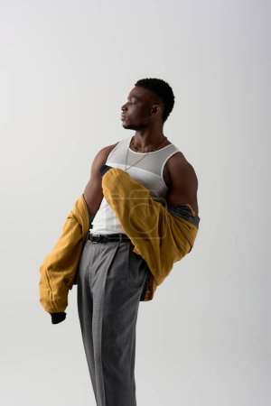 Photo for Side view of good looking afroamerican model in bomber jacket and sleeveless t-shirt posing isolated on grey, contemporary shoot featuring stylish attire, muscular - Royalty Free Image
