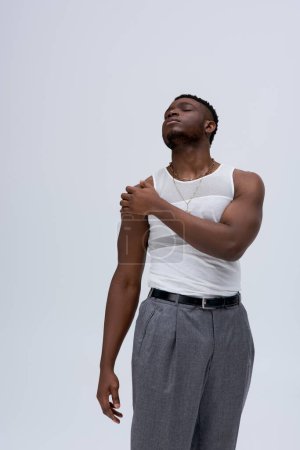 Low angle view of relaxed young afroamerican man in sleeveless t-shirt and pants touching shoulder isolated on grey, contemporary shoot featuring stylish attire, muscular