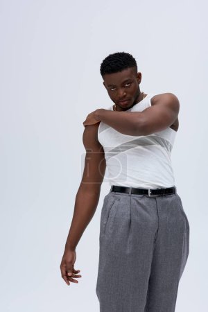 Confident and muscular african american man in sleeveless t-shirt and pants touching shoulder and looking at camera isolated on grey, contemporary shoot featuring stylish attire