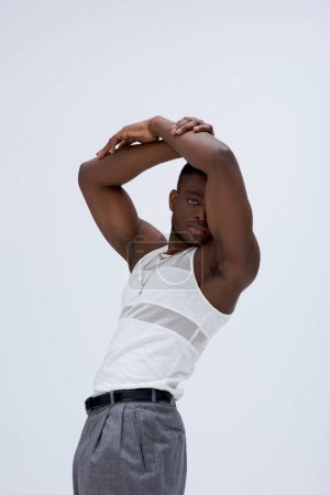 Muskulöser junger afroamerikanischer Mann in ärmellosem T-Shirt und Hose, der isoliert auf grauem, zeitgenössischem Shooting in stylischer Kleidung posiert und in die Kamera blickt