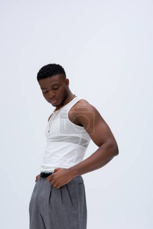 Fashionable and muscular young african american man in tank top and pants posing confidently in stylish and trendy outfit isolated on grey, good looking