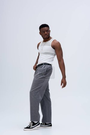 In voller Länge starker afroamerikanischer Mann in Hose und Tank-Top stehend und selbstbewusst posierend in stilvollem und trendigem Outfit auf grauem Hintergrund 
