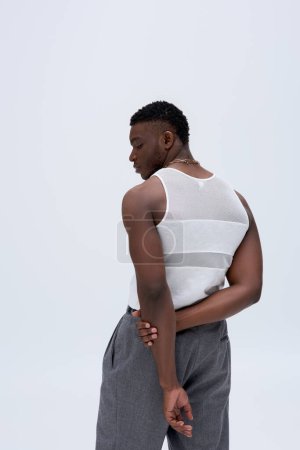 Téléchargez les photos : Vue latérale du jeune homme musclé afro-américain en pantalon et t-shirt sans manches posant en toute confiance dans une tenue élégante et tendance isolée sur gris, belle - en image libre de droit