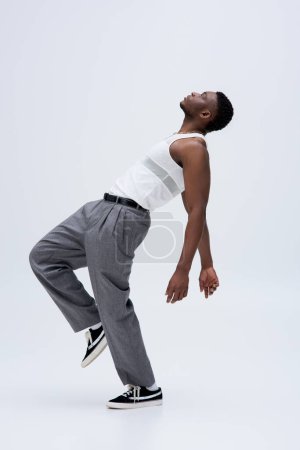 Side view of muscular afroamerican model in tank top and pants posing confidently in stylish and trendy outfit on grey background, good looking