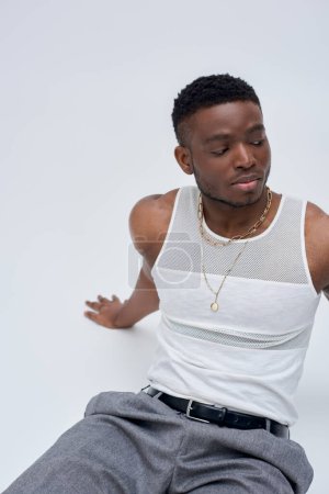 Good looking and young afroamerican man in tank top and pants sitting while posing confidently in stylish and trendy outfit on grey background, street style