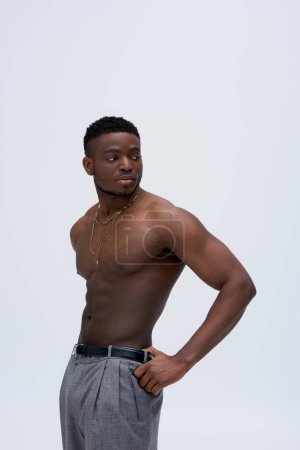 Muskulöser und sexy junger afroamerikanischer Mann in Halsketten und Hosen, isoliert auf grauen, selbstbewussten und modernen Posen stehend, Modeshooting, gut aussehend, Sinn für Mode