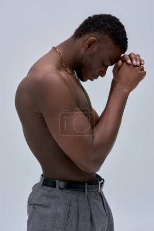 Side view of muscular and shirtless african american man in pants and golden necklaces standing isolated on grey, confident and modern pose, fashion shoot