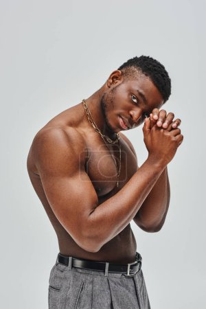 Muscular and good looking afroamerican man in golden necklaces and pants looking at camera while standing isolated on grey, confident and modern pose, fashion shoot