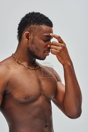 Shirtless and good looking african american model in golden necklaces touching nose isolated on grey, confident and modern pose, fashion shoot