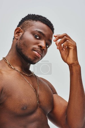 Portrait of muscular afroamerican man with golden necklaces looking at camera while standing isolated on grey, confident and modern pose, fashion shoot
