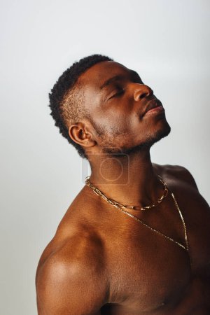 Homme afro-américain détendu et torse nu avec colliers dorés fermant les yeux isolés sur la pose grise, confiante et moderne, prise de vue mode