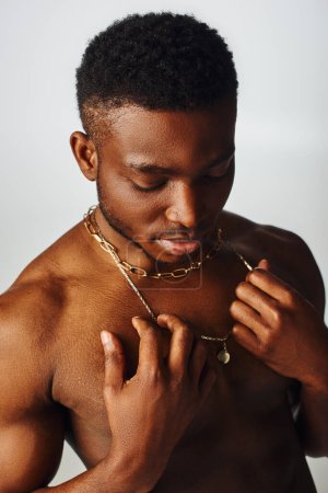 Jeune et torse nu afro-américain modèle masculin touchant colliers d'or et debout isolé sur la pose grise, confiante et moderne, tournage de mode