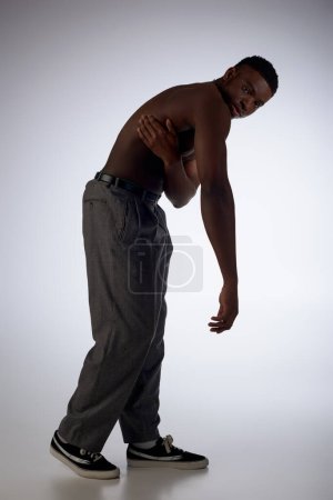 Volle Länge muskulöses und junges afrikanisch-amerikanisches Model in Hosen, das in die Kamera auf grauem Hintergrund blickt, selbstbewusst und modern posiert, Modeshooting, hemdloser Mann