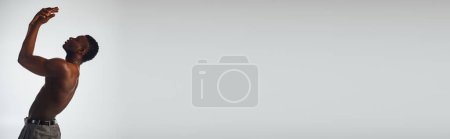 Téléchargez les photos : Vue latérale du modèle afroaméricain musclé et torse nu en pantalon isolé sur pose grise, confiante et moderne, prise de vue mode, bannière, homme torse nu - en image libre de droit