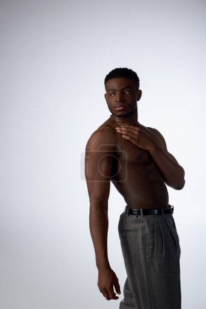 Hombre afroamericano sin camisa y joven en collares dorados y pantalones de pie en sombra aislado en pose gris, segura y moderna, sesión de moda, modelo muscular 