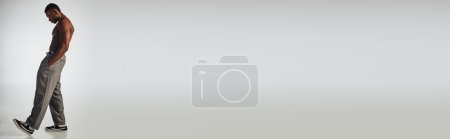 Téléchargez les photos : Longueur totale du modèle afroaméricain torse nu en colliers et pantalons dorés tenant la main dans la poche sur fond gris, pose confiante et moderne, prise de vue mode, bannière - en image libre de droit