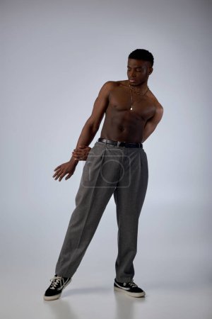 Longitud completa de modelo afroamericano muscular en collares dorados, pantalones y zapatillas de deporte de pie sobre fondo gris, pose segura y moderna, sesión de moda, hombre sin camisa