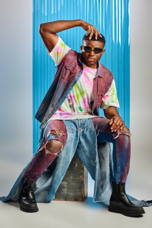 Photo for Stylish afroamerican man in sunglasses, denim vest and colorful t-shirt touching head while sitting on stone on grey with blue polycarbonate sheet at background, fashion shoot, sustainable fashion - Royalty Free Image