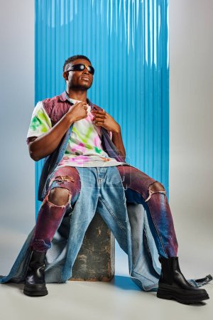 Voller Länge trendiges afrikanisch-amerikanisches Model in Sonnenbrille, farbenfroher Jeansweste und T-Shirt sitzend auf Stein auf Grau mit blauem Polycarbonat-Laken im Hintergrund, Mode-Shooting, DIY-Kleidung