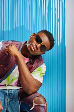 Trendiger junger afroamerikanischer Mann mit Sonnenbrille, Jeansweste und zerrissenen Jeans posiert auf grau mit blauem Polycarbonat-Laken im Hintergrund, Mode-Shooting, nachhaltige Mode, DIY-Kleidung