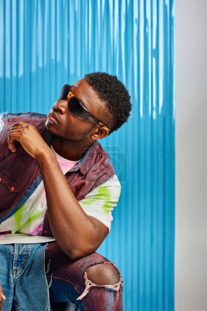 Modelo afroamericano joven y elegante en camiseta colorida, gafas de sol y jeans rasgados posando en gris con sábana de policarbonato azul en el fondo, moda, estilo de vida sostenible, ropa de bricolaje