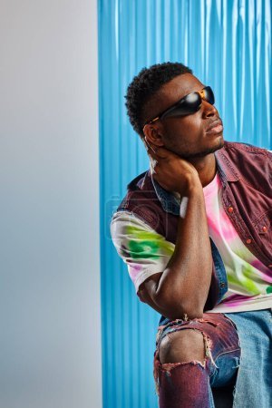 Trendy young afroamerican man in sunglasses, colorful t-shirt and ripped jeans touching neck and posing on grey with blue polycarbonate sheet at background, fashion shoot, DIY clothing