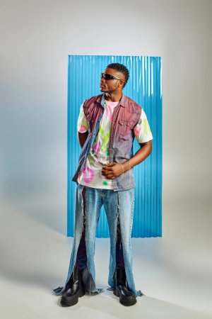 Voller Länge trendiges afrikanisch-amerikanisches Model in farbenfrohem T-Shirt, Sonnenbrille und zerrissenen Jeans, stehend auf grau mit blauem Polycarbonat-Laken im Hintergrund, Mode-Shooting, nachhaltiger Lebensstil