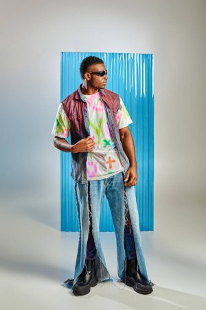 Pleine longueur de beau et jeune homme afro-américain en lunettes de soleil, gilet en denim et jeans déchiré debout sur gris avec feuille de polycarbonate bleu à l'arrière-plan, tournage de mode, vêtements de bricolage