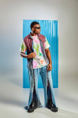 Full length of good looking and young afroamerican man in sunglasses, denim vest and ripped jeans standing on grey with blue polycarbonate sheet at background, fashion shoot, DIY clothing mug #658610162