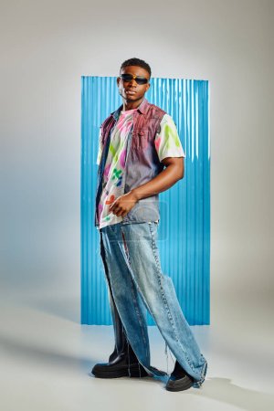Mannequin afro-américain jeune et élégant en lunettes de soleil, jean déchiré et gilet en denim sur fond gris avec une feuille de polycarbonate bleu, prise de vue mode, vêtements DIY