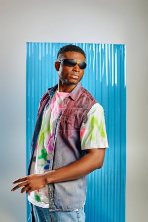 Homme afro-américain à la mode en lunettes de soleil, gilet en denim coloré et t-shirt posant et debout sur gris avec une feuille de polycarbonate bleu à l'arrière-plan, mode durable, vêtements DIY
