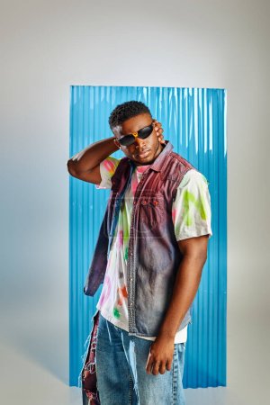 Selbstbewusster junger afroamerikanischer Mann mit Sonnenbrille, buntem T-Shirt und Jeansweste, der den Kopf berührt und auf grau mit blauem Polycarbonat-Laken im Hintergrund steht, nachhaltige Mode, DIY-Kleidung