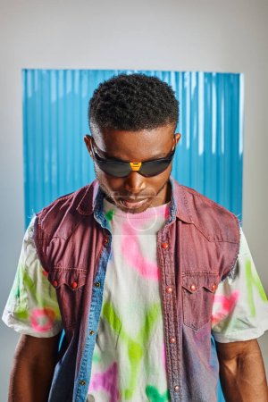Portrait of young and stylish afroamerican man with trendy hairstyle wearing sunglasses, denim vest and colorful t-shirt on grey with blue polycarbonate sheet at background, sustainable fashion