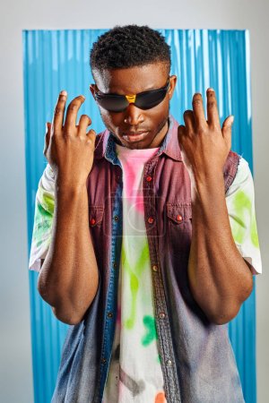 Portrait of stylish young afroamerican model in sunglasses, colorful denim vest and t-shirt posing and standing on grey with blue polycarbonate sheet at background, sustainable fashion, DIY clothing