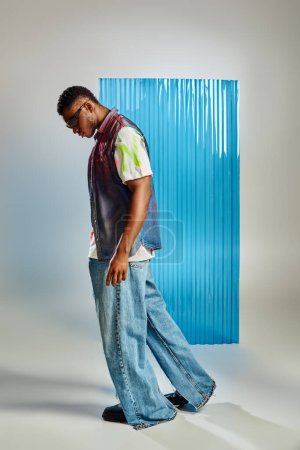 Foto de Vista lateral del modelo afroamericano de moda en gafas de sol, chaleco vaquero y jeans caminando sobre gris con lámina de policarbonato azul de fondo, moda sostenible, ropa de bricolaje - Imagen libre de derechos