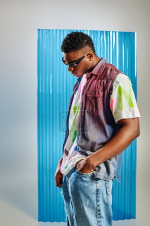 Hombre afroamericano joven de moda en gafas de sol, camiseta colorida y chaleco de mezclilla tomados de la mano en el bolsillo de vaqueros rasgados en gris con sábana de policarbonato azul en el fondo, ropa de bricolaje