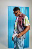 Trendy young afroamerican man in sunglasses, colorful t-shirt and denim vest holding hand in pocket of ripped jeans on grey with blue polycarbonate sheet at background, DIY clothing Stickers #658610350