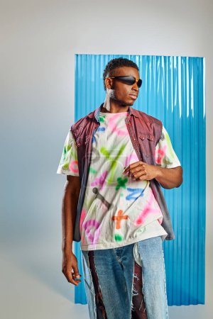 Fashionable young afroamerican model in sunglasses, colorful denim vest and ripped jeans standing on grey with blue polycarbonate sheet at background, sustainable fashion, DIY clothing