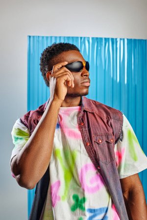 Portrait of trendy afroamerican man touching sunglasses while wearing denim vest and colorful t-shirt on grey with blue polycarbonate sheet at background, sustainable fashion, DIY clothing