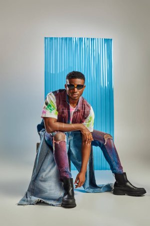 afroamerican model in sunglasses, colorful denim vest and contemporary ripped jeans sitting on stone on grey with blue polycarbonate sheet at background, sustainable fashion, DIY clothing