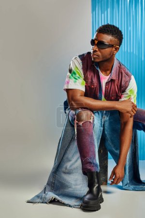 Homme afro-américain confiant en lunettes de soleil, gilet en denim et jeans déchiré tendance assis sur pierre sur fond gris avec une feuille de polycarbonate bleu, mode durable, vêtements de bricolage
