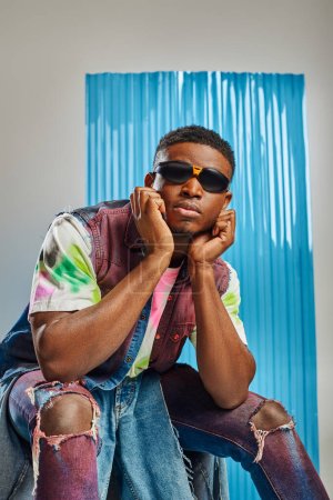 Portrait of young afroamerican model in sunglasses and colorful denim vest and ripped jeans sitting on grey with blue polycarbonate sheet at background, sustainable fashion, DIY clothing