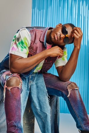 Téléchargez les photos : Jeune modèle afro-américain à la mode en lunettes de soleil, jean déchiré coloré et gilet en denim assis sur pierre sur gris avec une feuille de polycarbonate bleu à l'arrière-plan, tournage de mode, vêtements DIY - en image libre de droit