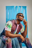 Confident afroamerican man in colorful ripped jeans, sunglasses and denim vest sitting on grey with blue polycarbonate sheet at background, fashion shoot, DIY clothing, sustainable fashion Mouse Pad 658610492