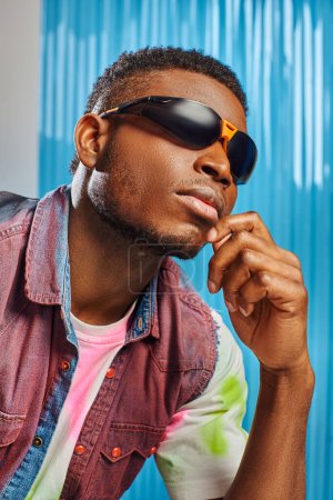 Portrait of young afroamerican man with trendy hairstyle, sunglasses and colorful denim vest posing on grey with blue polycarbonate sheet at background, fashion shoot, DIY clothing 
